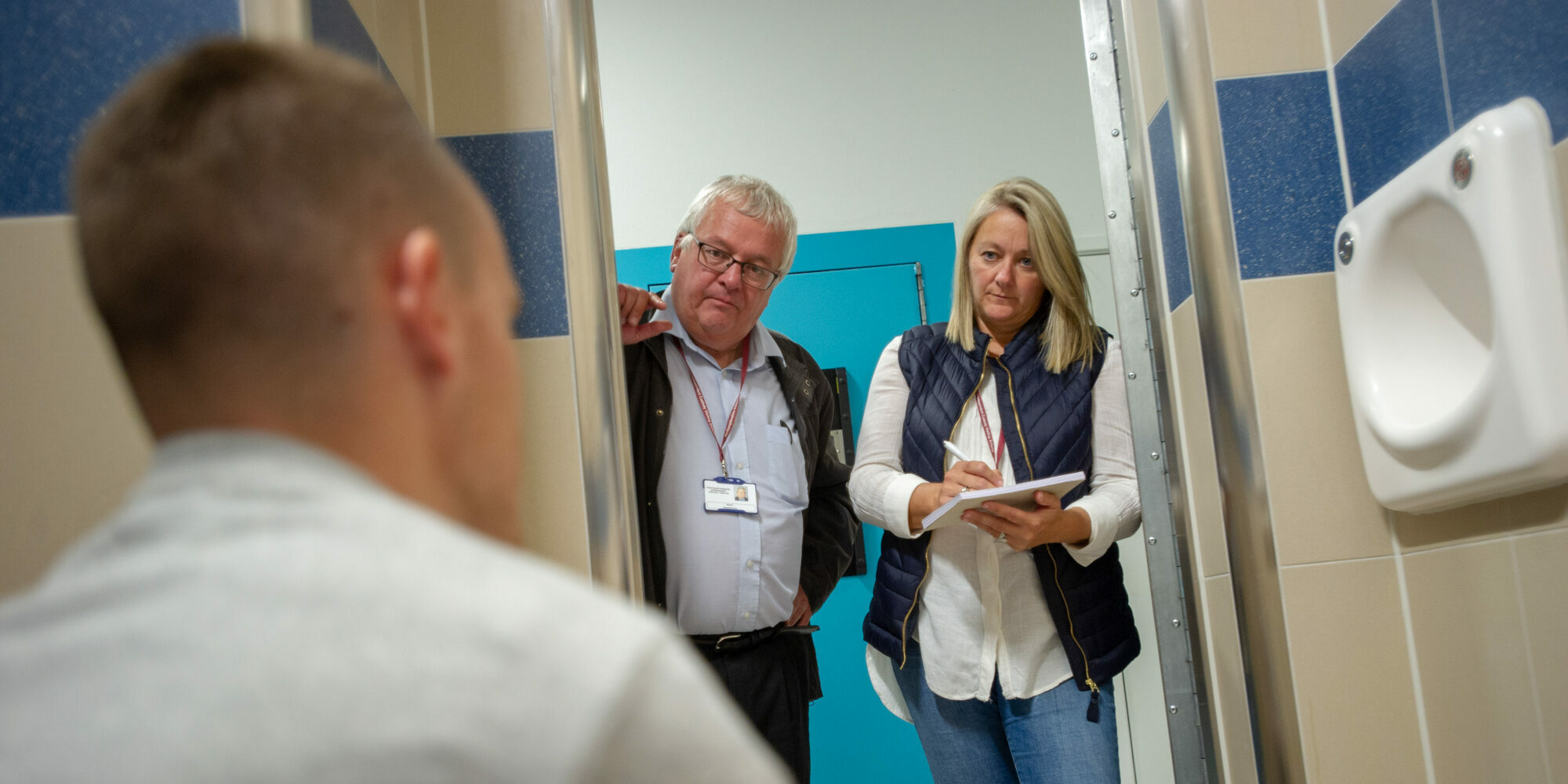Two volunteers for the Independent Custody Visiting (ICV) scheme are pictured in a custody cell speaking to a detainee, who is not fully identified