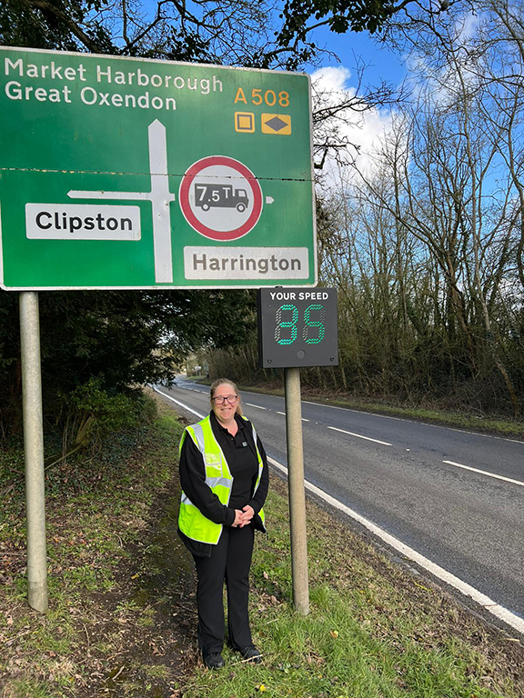 A lady wearing a high-vis vest standing next to a road and road sign