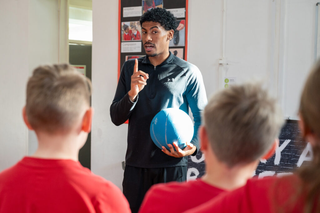 Andre Arissol holding a blue basketball speaking to a group of school children