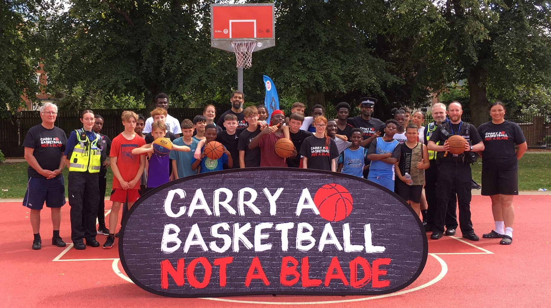 A group of adults and children including police officers and community support officers, outside on a sports court. Some are wearing sports attire, some are holding basketballs. They are in front of a basketball hoop and new and behind a large sign saying 