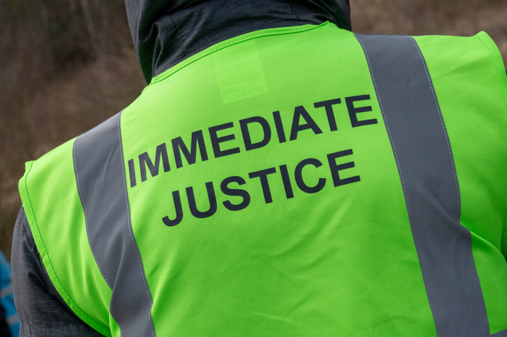 The back of a person is shown, they are wearing a green vest with the words Immediate Justice printed on them