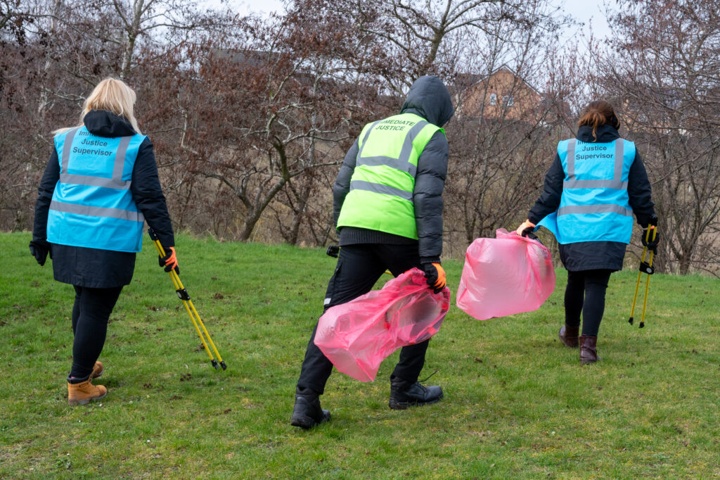 An offender, wearing a green vest, is accompanied by two Immediate Justice Supervisors either side wearing blue vests. They are holding pink rubbish bags during a litter pick.
