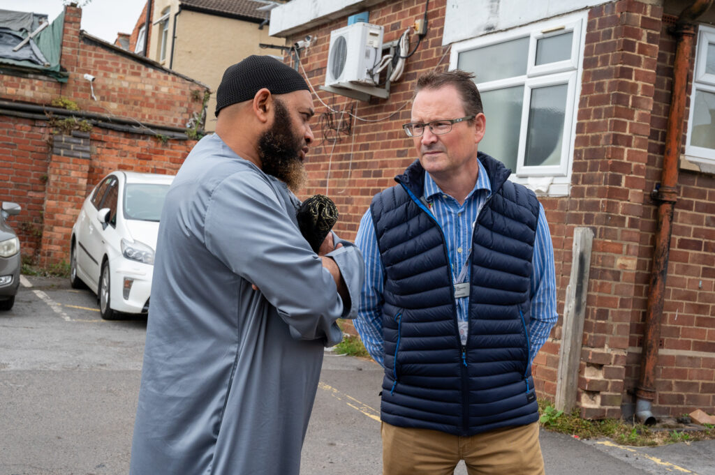 Councillor David Smith wearing navy gilet over a blue striped shirt, talking to the Imam from Clare Street Mosque wearing traditional wear