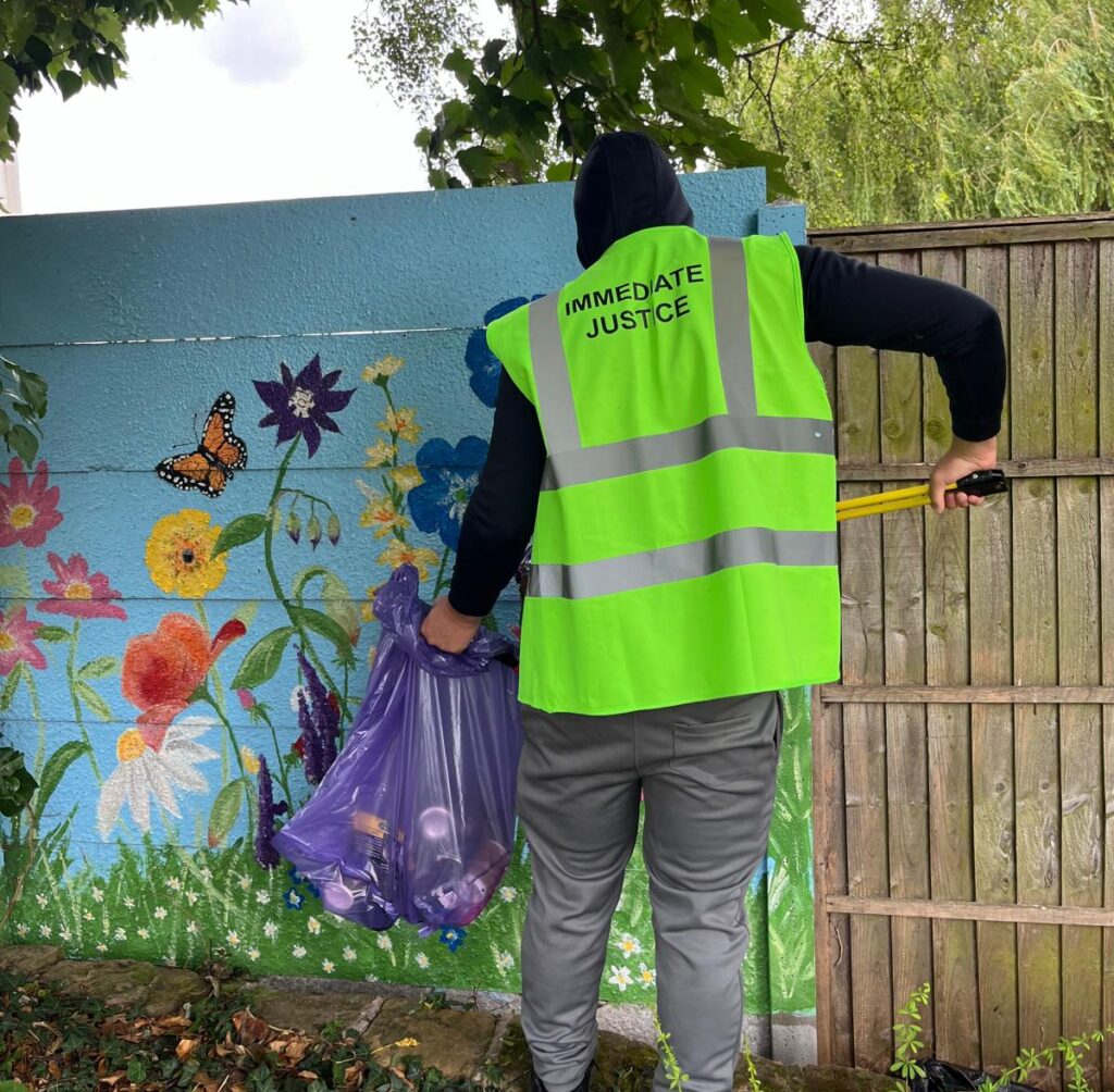 A person wearing a yellow high visibility vest picks up litter in front of a mural in a park