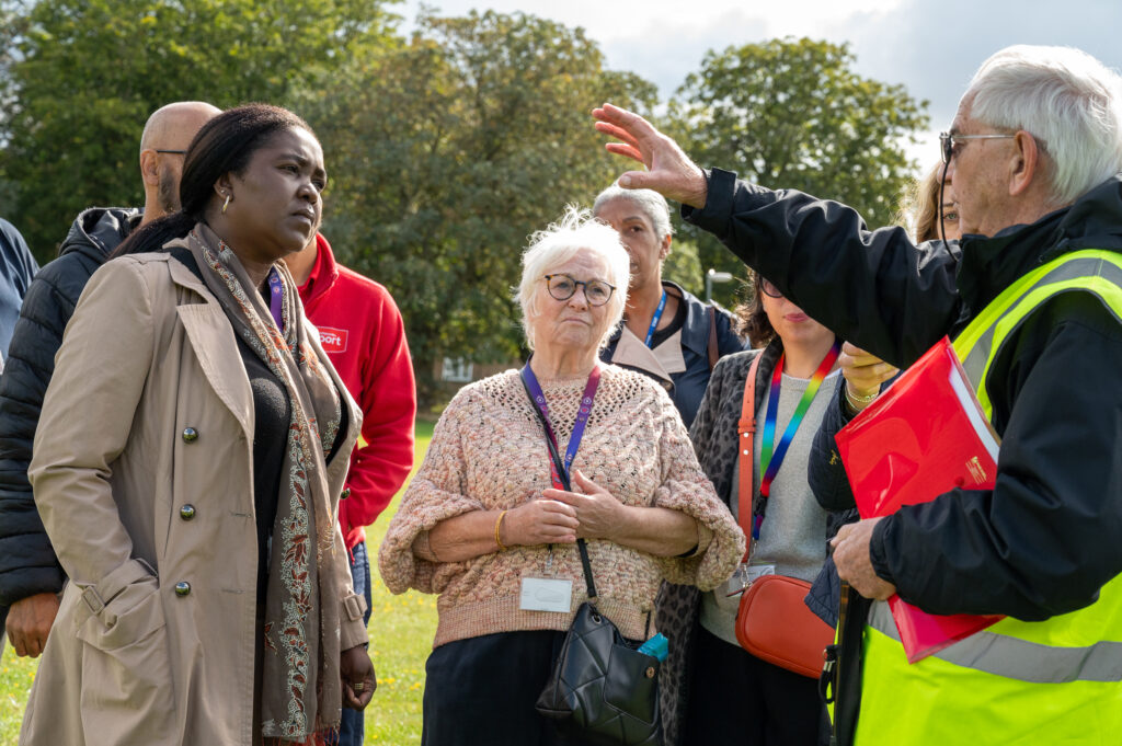 Danielle Stone wearing a light pink jumper stands in the middle of a group of people wearing hi-visibility jackets, standing next to her is Marianne Kimani wearing a long tan coat