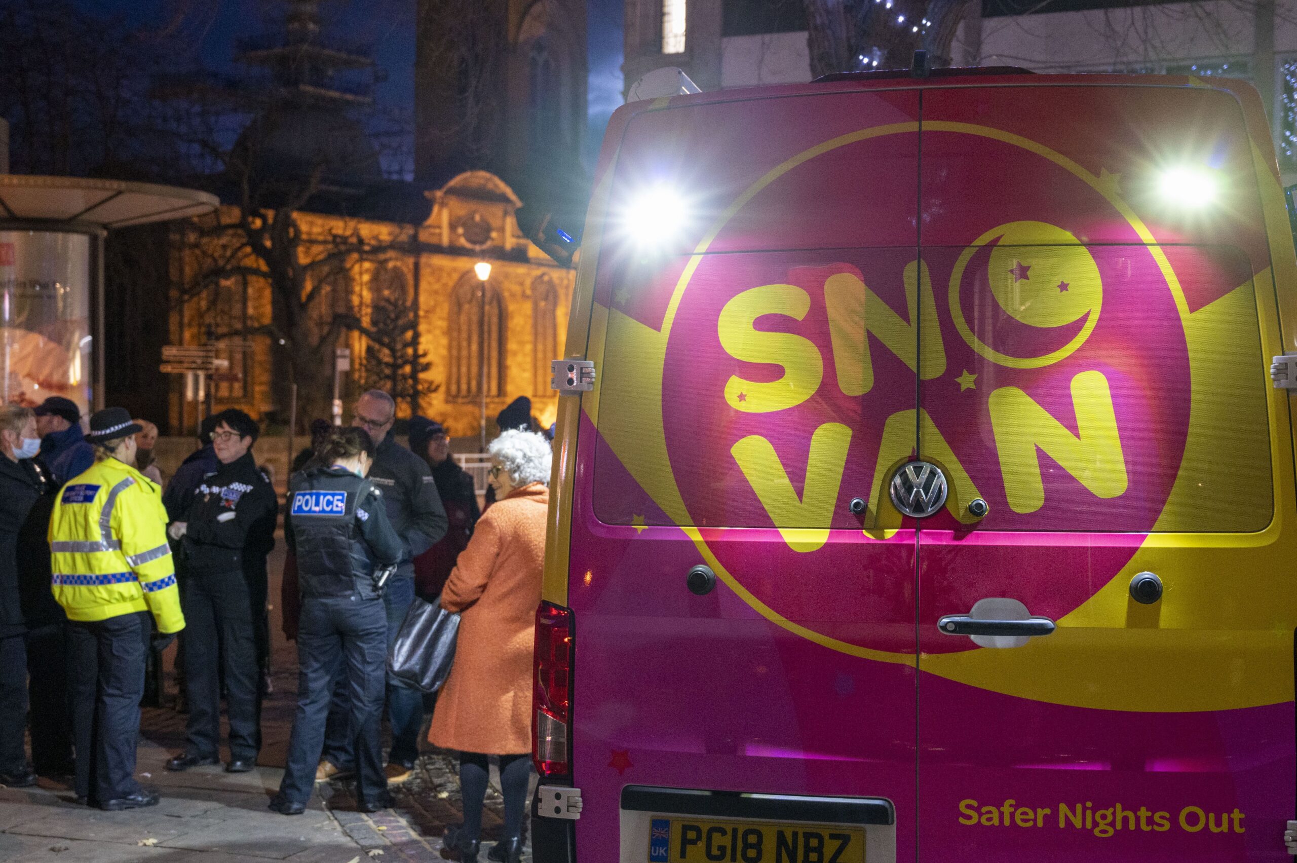 The back of a Pink van, that has a large yellow SNO VAN logo on it. Surrounded by members of the pubic and police officers in uniform.