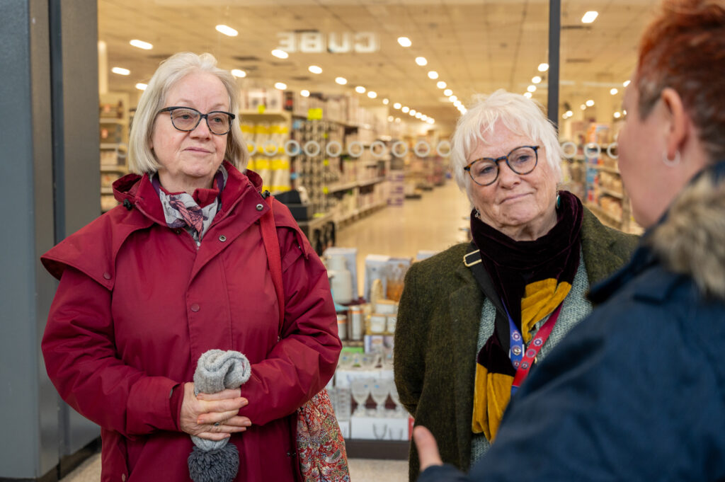 Deputy Mayor of Wellingborough, Councillor Viv Wilkinson, is shown on the left in a red coat. Alongside her, wearing a green coat and black scarf, is Police, Fire and Crime Commissioner Danielle Stone. They are shown speaking to a Community Safety Officer, who is blurred on the right of the picture.