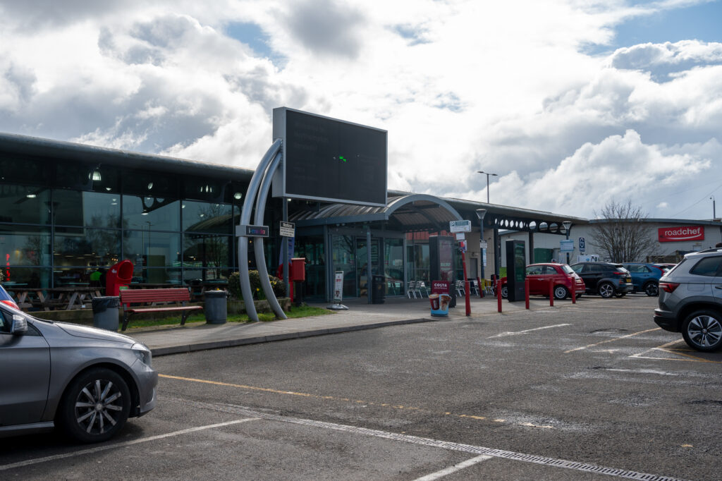 A picture shows the forecourt of the Roadchef Northampton service station. A number of cars are shown, and there is a big black screen in the centre of the picture near the entrance door to the building.