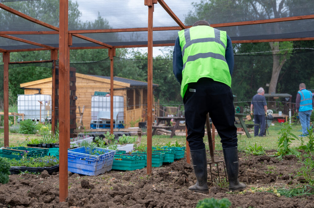  A man wearing black trousers and a green hi-vis is gardening into mud at an allotment. In the background there is a person wearing a green t-shirt and another person wearing a blue hi-vis.