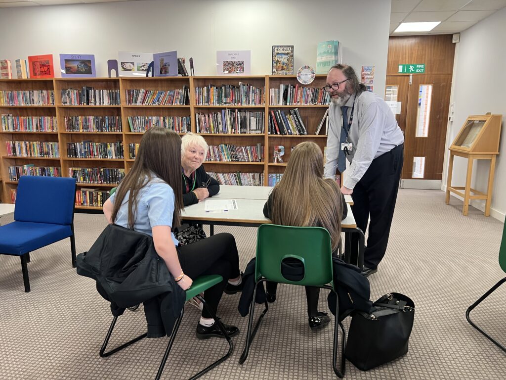 Danielle Stone sits at a white table wearing a black top, she is talking to two young girls with long brown hair, wearing blue shirts. A man with dark hair is standing over the table wearing a long sleeve pale blue shirt.