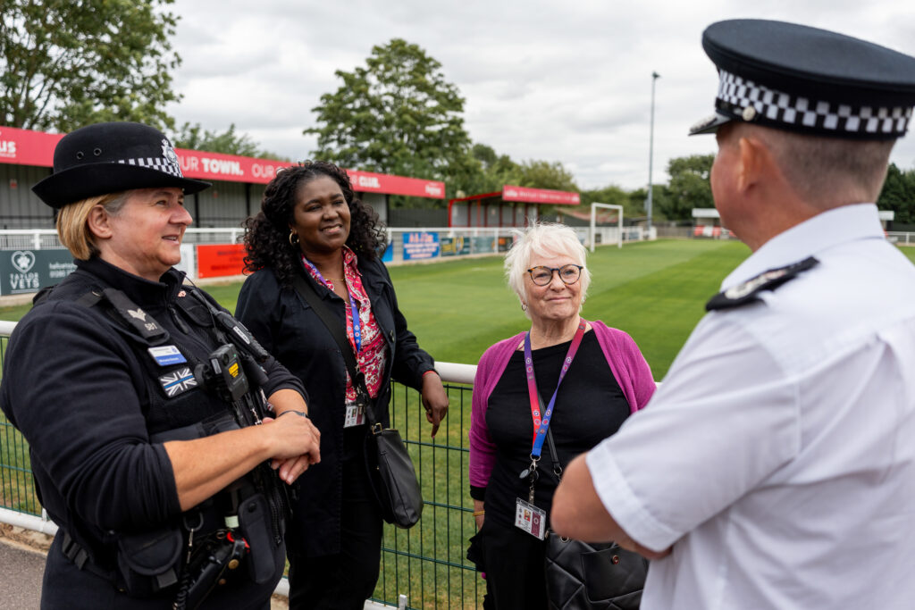 One female police officer wearing all black uniform and a hat, one woman wears a pink top and a black jacket, one woman in shorter with white hair wears a black dress and a purple cardigan, and one man, with his back to the camera, wears a white police uniform and a black hat.
They sit around talking