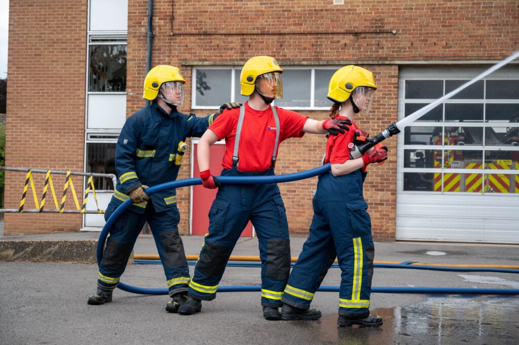 three young people, two wearing dark blue trousers and red t-shirts, one is wearing all dark blue. All three are wearing yellow helmets. Together they are holding a hose-reel with water shooting out of it.