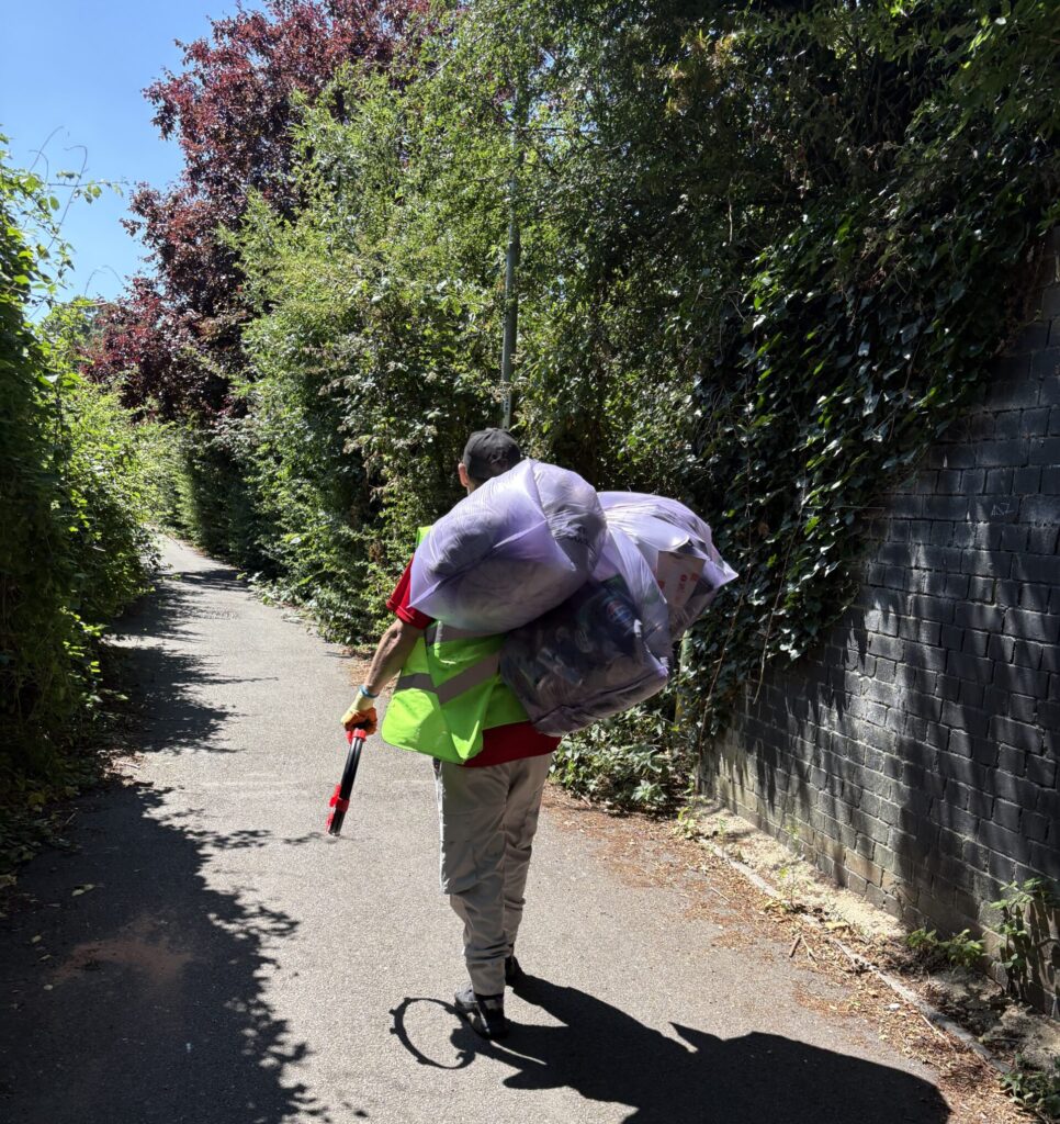 A man walks down a path wearing pale trousers and a yellow hi-vis, he is carrying multiple bags of rubbish over his shoulder
