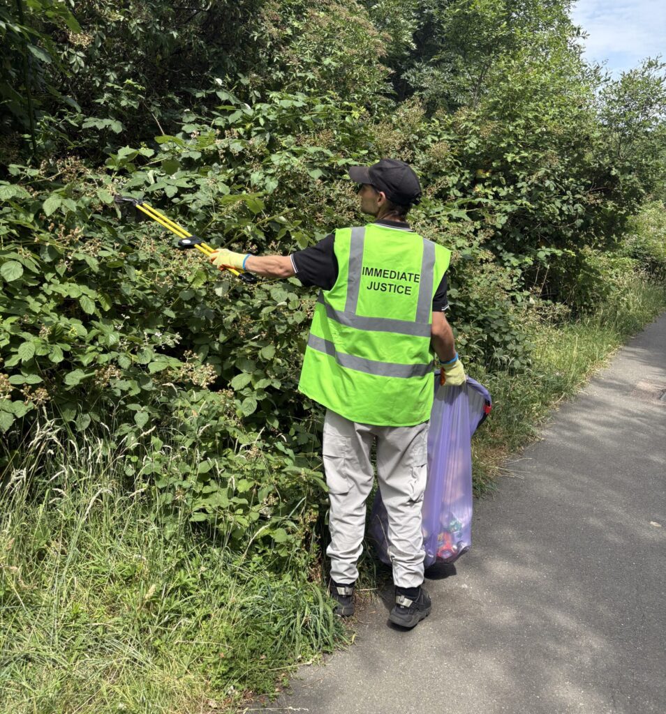 A man carrying a purple bin bag and a litter picker picks rubbish out of a bush.

He is wearing pale grey trousers and a yellow hi-vis vest