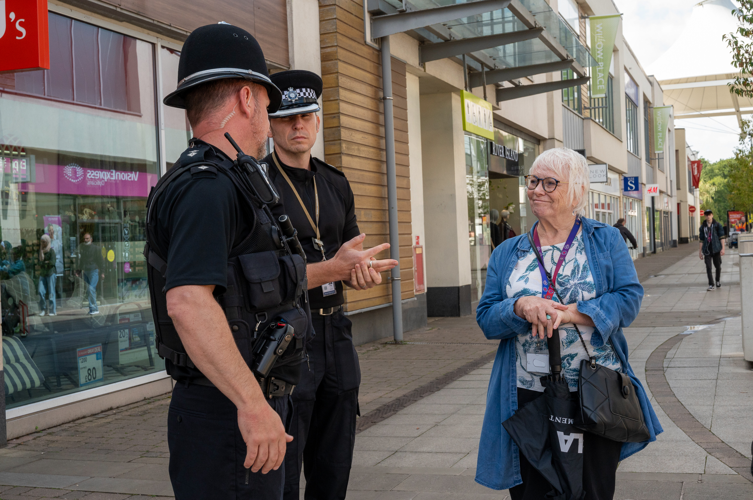 Two police officers in uniform are standing on a pedestrian street, engaging in conversation with an older woman who is wearing a blue jacket and holding a black bag. The background shows various storefronts and people walking along the street. The faces of the individuals are blurred for privacy.