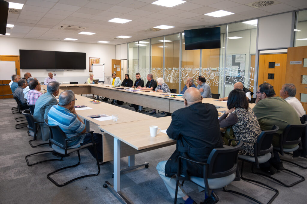 A group of people are seated around a large U-shaped conference table in a meeting room. The room has glass walls on one side, with doors and windows visible. Two large flat-screen monitors are mounted on the front wall, along with a whiteboard. The attendees appear to be engaged in discussion, with some taking notes or reviewing documents. The ceiling features recessed lighting, and the tables hold various office supplies such as cups, papers, and pens.