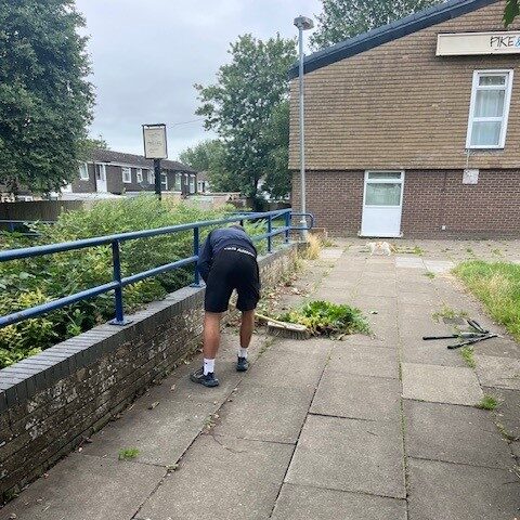 A person wearing a black shirt, black shorts, white socks, and black shoes is bending over near a blue railing on a paved walkway, appearing to remove weeds or plants. Tools and plant debris are scattered nearby. In the background, a brick building with a partially visible sign reading "PIKE" is surrounded by trees and bushes.