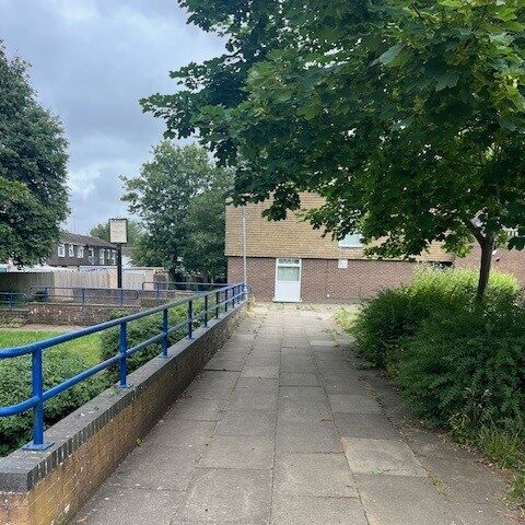 A paved walkway leads to a brick building with a white door. A blue handrail runs along the left side of the path, flanked by trees with green foliage. The sky is cloudy, and additional buildings are visible in the background.