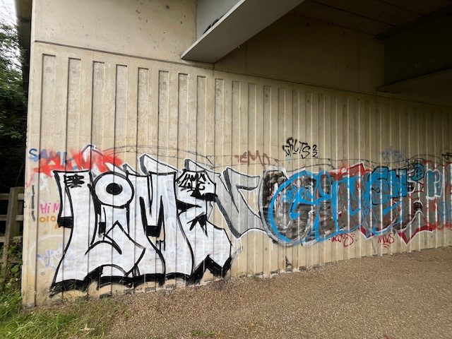  A concrete wall under a bridge covered with multiple graffiti tags. Prominent graffiti includes 'LIME' in white with black outlines and 'GAMER' in blue with black outlines, surrounded by smaller tags and scribbles. The ground is paved with a gravel path leading to the wall, with some grass visible in the bottom left corner.