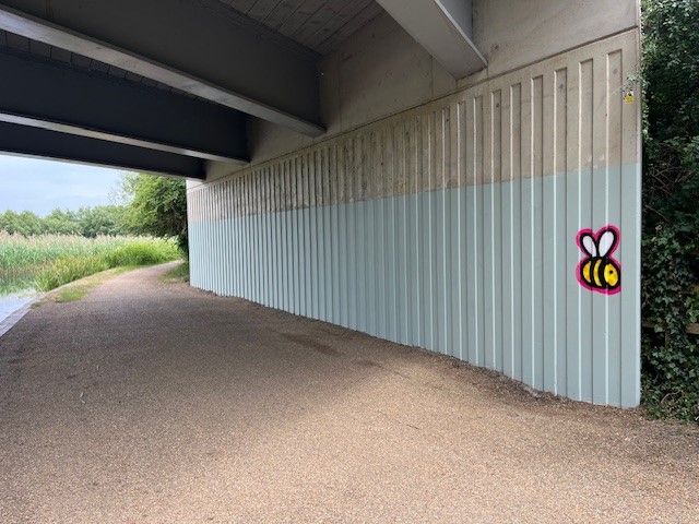 A pathway under a bridge with a corrugated metal wall on the right, painted in two shades of gray. A cartoon-style bee graffiti is visible on the wall near some greenery. The path curves to the left, bordered by tall grass and bushes on the left side.