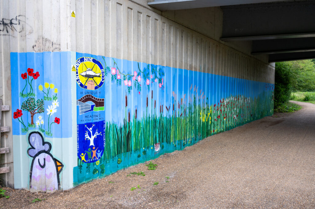 A mural painted on the side of an underpass, depicting a vibrant nature scene with flowers, plants, and trees. On the left, a cartoon-style chicken head with closed eyes and a small smile is surrounded by red flowers and green leaves. The center features two circular logos: one with a yellow sun and the word 'SUNSHINE,' and another with white deer antlers on a blue background labeled 'MEADOWS.' The mural continues with more greenery and colourful flowers extending along the wall.