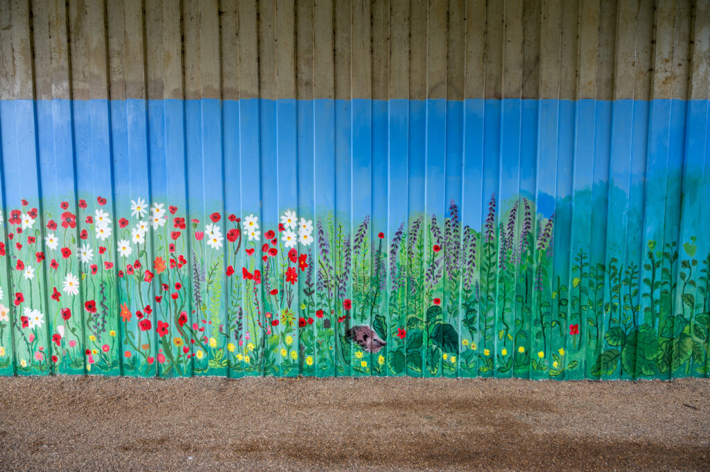 A vibrant mural painted on a corrugated metal wall, depicting a meadow scene with green plants, red poppies, white daisies, yellow blossoms, and taller purple flowers. The background is shaded blue to represent the sky, and the ground in front of the mural is covered with gravel or small pebbles.