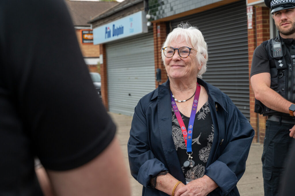 A person with white hair, wearing a dark jacket and a lanyard with various badges, stands outside in front of a shop. Two other individuals are partially visible; one on the left side of the image and another on the right, who appears to be a police officer based on the uniform and equipment. The background includes brick buildings and another shop sign