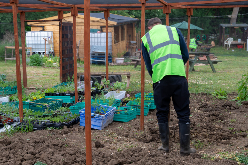 A person wearing a bright green safety vest and black rubber boots is working in a garden area. They are standing on freshly tilled soil, surrounded by plastic crates filled with plants and seedlings. In the background, there are wooden structures like a shed and other gardening equipment, with greenery visible in the distance.