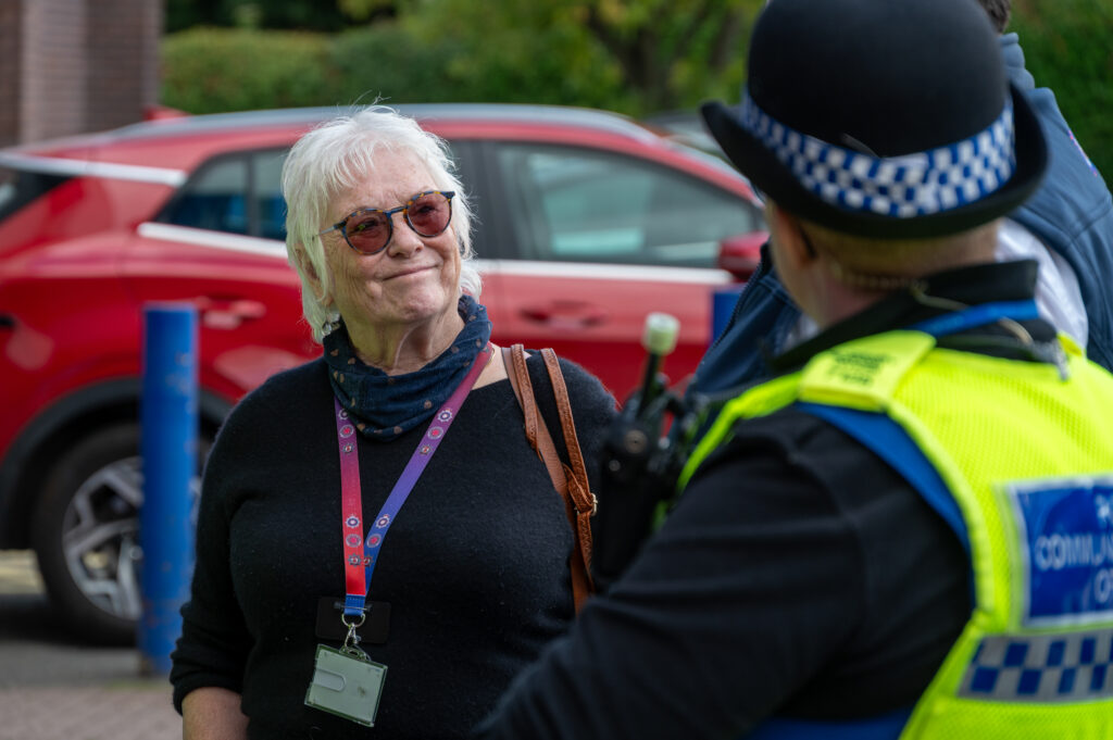 Two individuals interacting outdoors: one wearing a black top, glasses, and a lanyard with an ID card, and the other in a high-visibility police uniform and hat, with a red car and greenery in the background