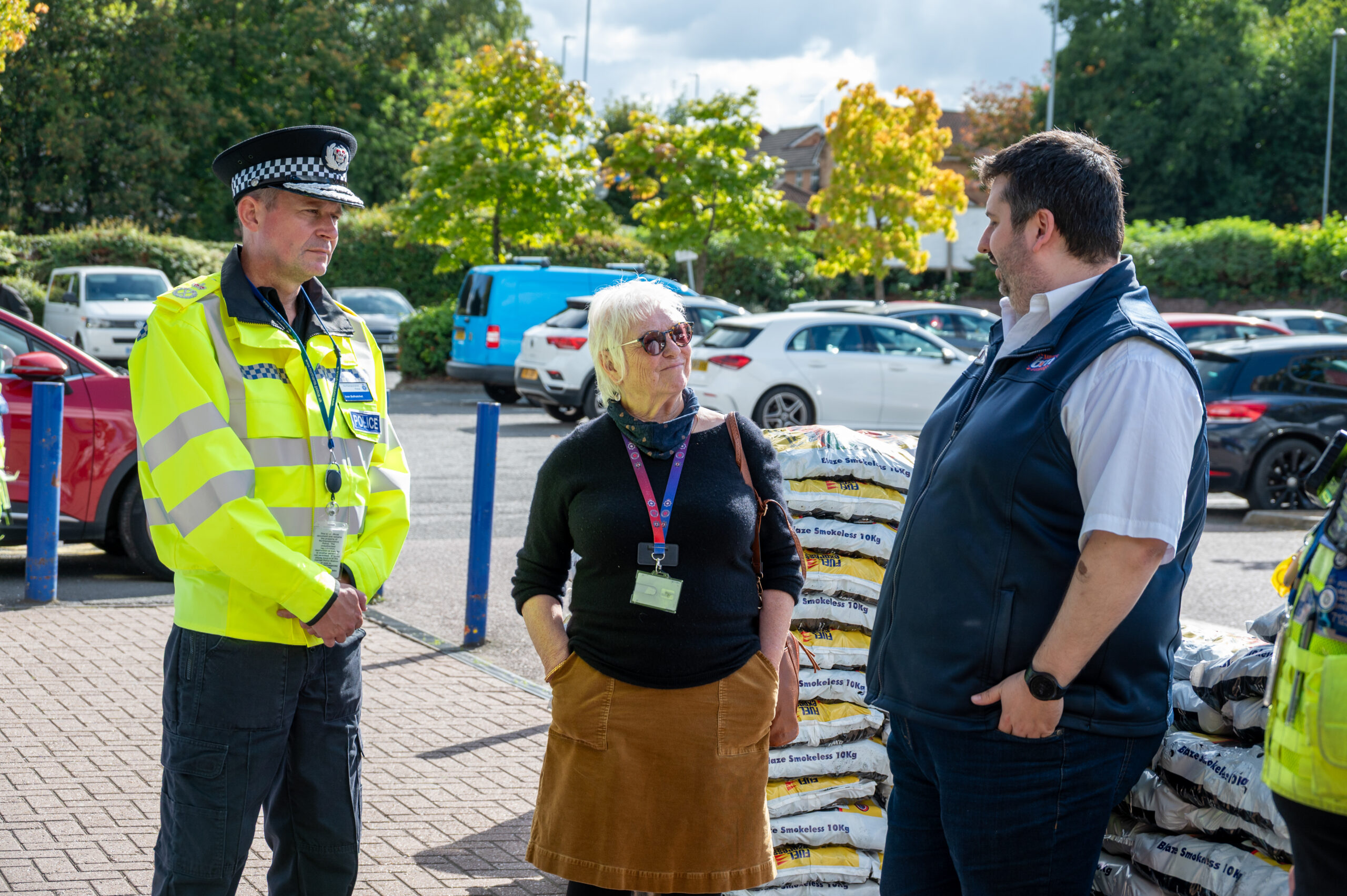 Three individuals standing in a parking lot during a community event: one in a police uniform, one in casual attire with an ID badge, and one in business casual clothing with a vest and ID badge, engaged in conversation with parked cars and autumn trees in the background