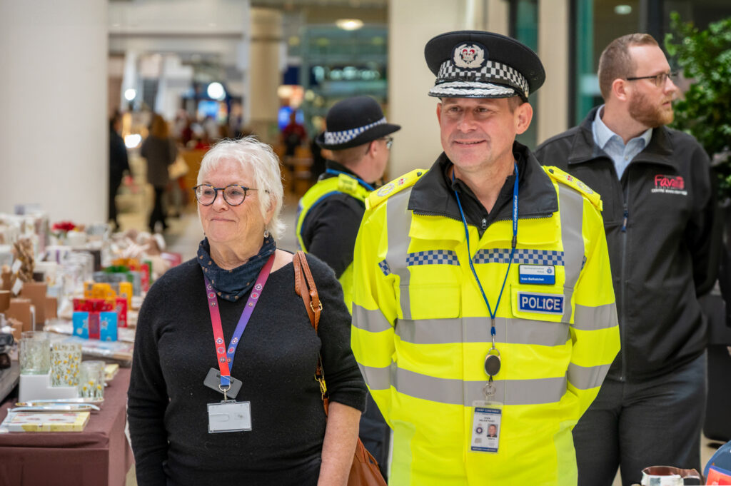 Two individuals in the foreground at a public indoor event: one wearing a black top with a lanyard and name badge, and the other in a high-visibility police uniform and hat. Behind them are tables with displayed items and other attendees, including another officer and a person in business attire