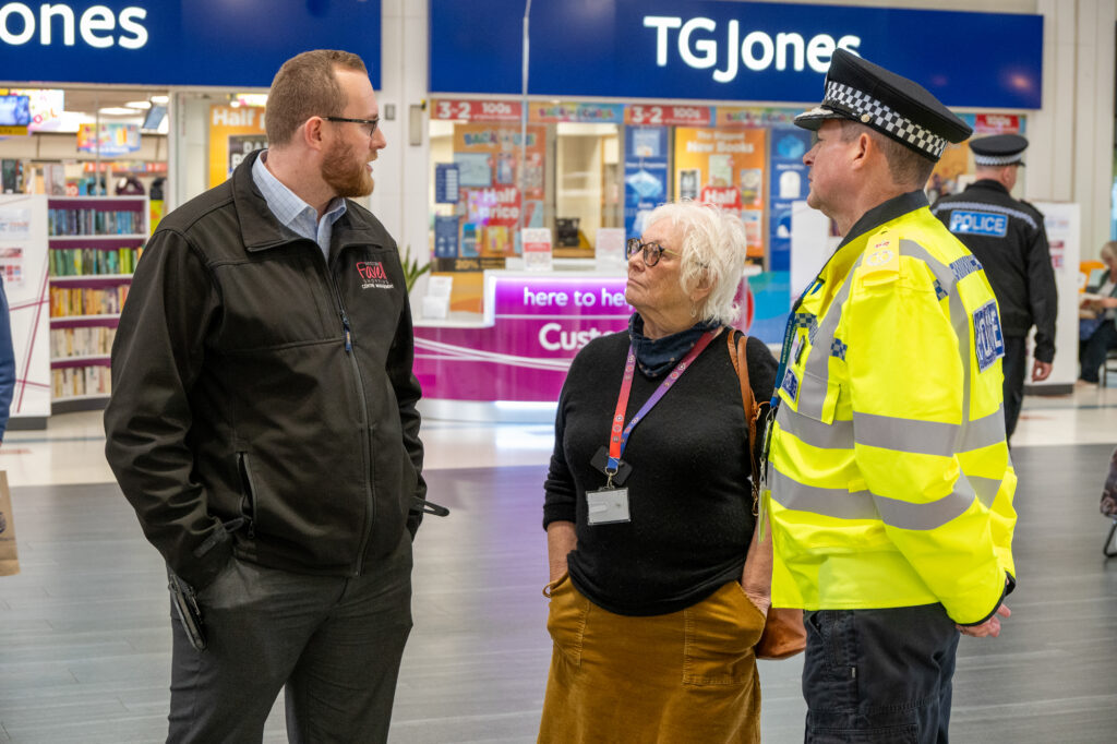 Three individuals conversing in a public indoor space, including a person in a TGJones-branded jacket, another in casual attire with a lanyard and badge, and a third in a high-visibility police uniform, with storefronts and promotional signage in the background