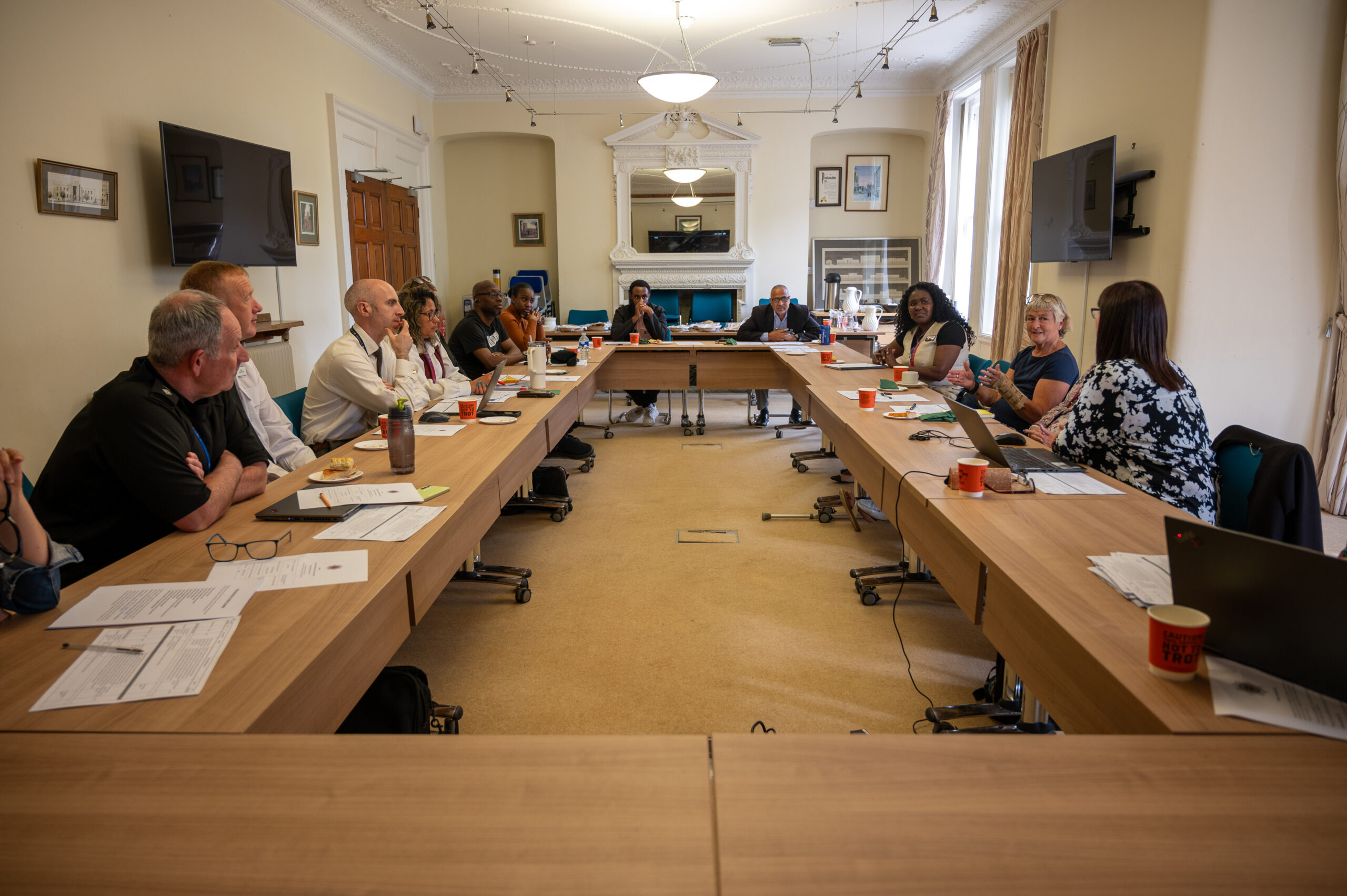 A group of people seated around a large U-shaped conference table in a well-lit meeting room, engaged in discussion. The table is covered with papers, laptops, and coffee cups. The room features beige walls, framed pictures, large windows with curtains, two wall-mounted flat-screen TVs, and a fireplace with a mirror above it.