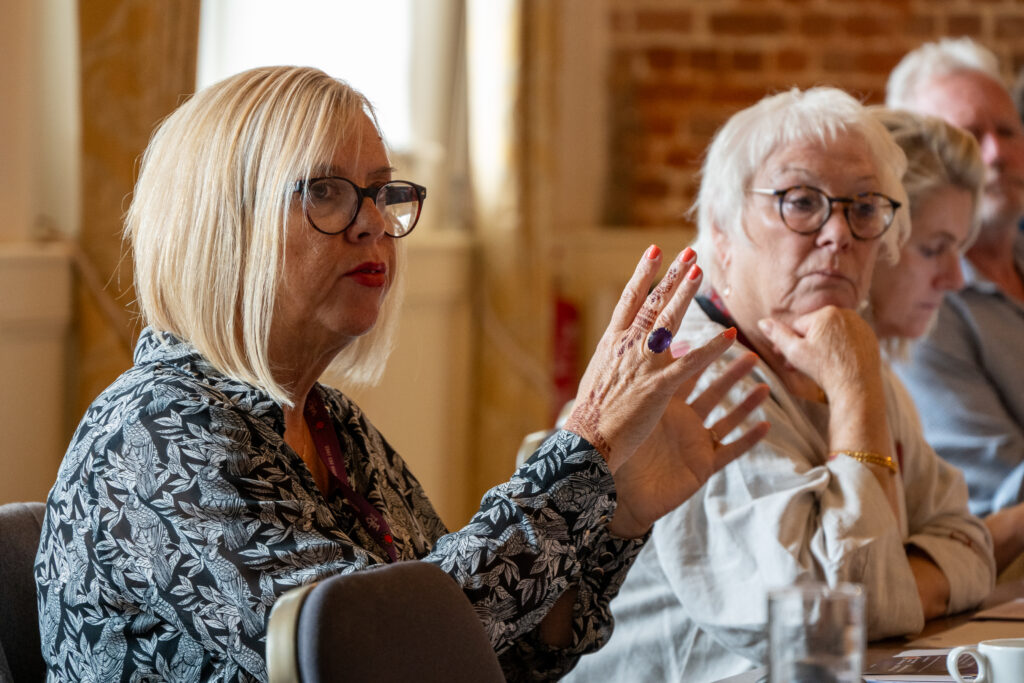 Two individuals are highlighted in a meeting; one (Fiona Campbell) speaks and gestures with both hands while the other (Danielle Stone) listens, with more participants in the background.