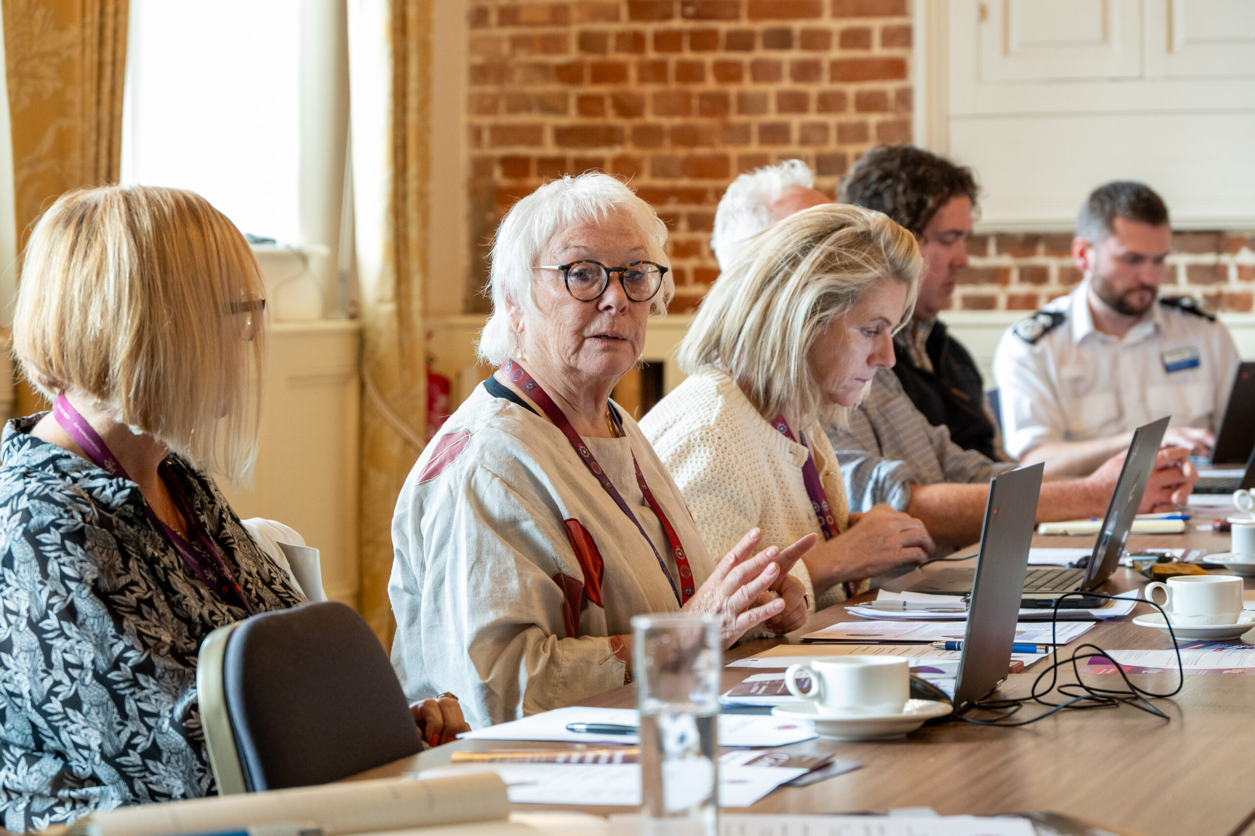 Several people seated around a table in a meeting, some using laptops, while one person in a white top - Police, Fire and Crime Commissioner Danielle Stone - is shown speaking and others listen attentively.