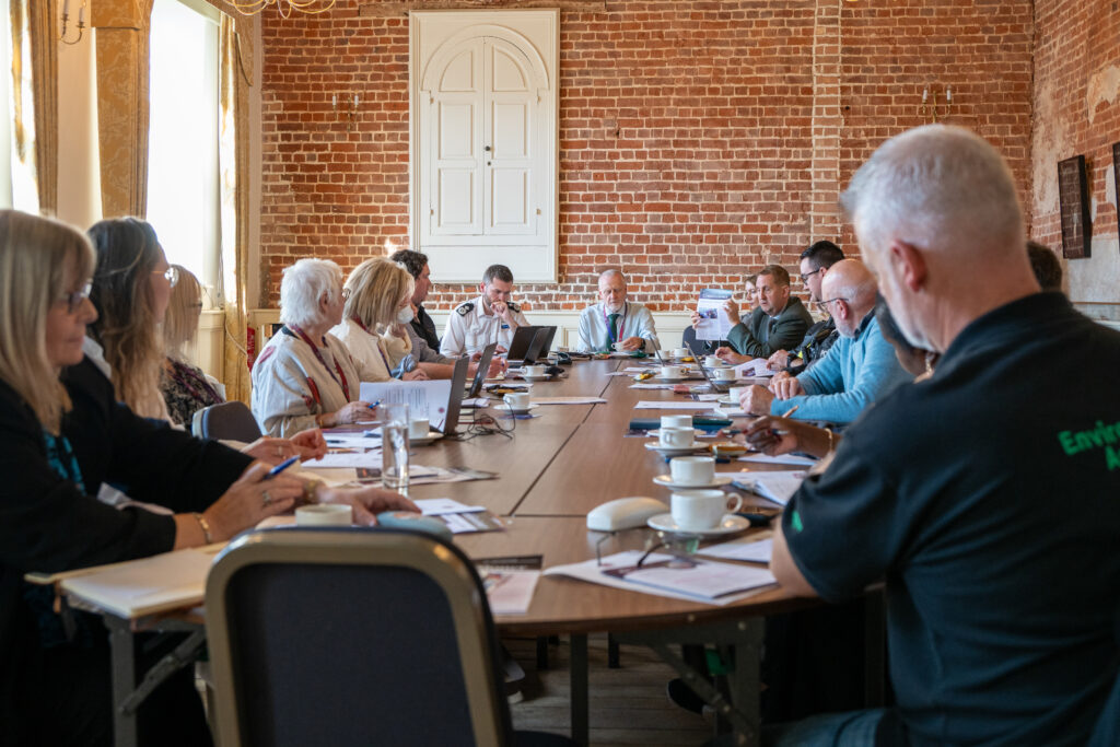 A wide view of a meeting room with large windows and brick walls, showing multiple people seated around a table engaged in discussion.