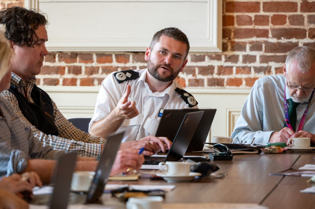 A person in uniform with epaulettes speaks and gestures during a meeting in a brick-walled room, using a laptop at a roundtable with other attendees.