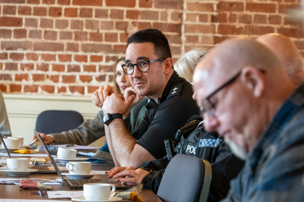A meeting scene showing participants focused on laptops and notes, with one person wearing glasses looking toward the camera.