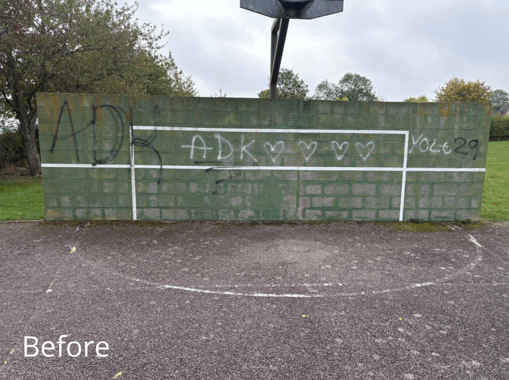 An outdoor sports wall with a dark green surface covered in graffiti, including large black letters and white hearts. White lines forming a rectangular goal outline are visible but partially obscured by graffiti. A basketball hoop is mounted above the wall. The ground in front shows faded court markings, and there are trees and grass in the background under an overcast sky. The word “Before” is written in white text at the bottom left corner.