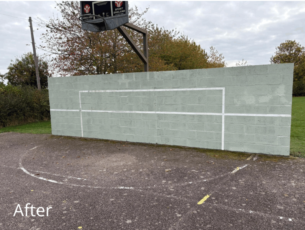 A freshly repainted outdoor sports wall with a light green surface and crisp white lines forming a rectangular goal outline. The wall is clean and free of graffiti. A basketball hoop with a Sport England logo is mounted above the wall. The ground in front shows faded court markings, and there are trees and grass in the background under a cloudy sky. The word “After” is written in white text at the bottom left corner.