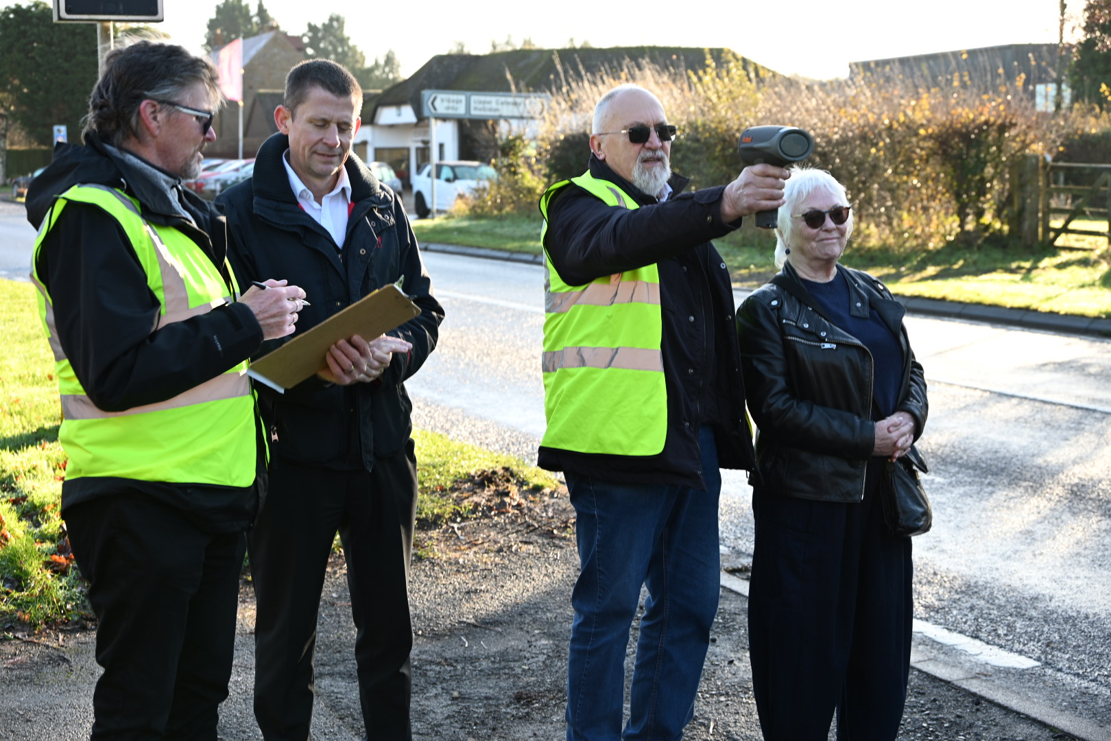 Four people standing on the roadside during daylight, two wearing high-visibility vests. One person is holding a clipboard and writing, while another is pointing a handheld speed detection device toward the road. The group appears to be engaged in a traffic monitoring or speed check activity. In the background, there are hedges, a building with signage, and a clear sky.