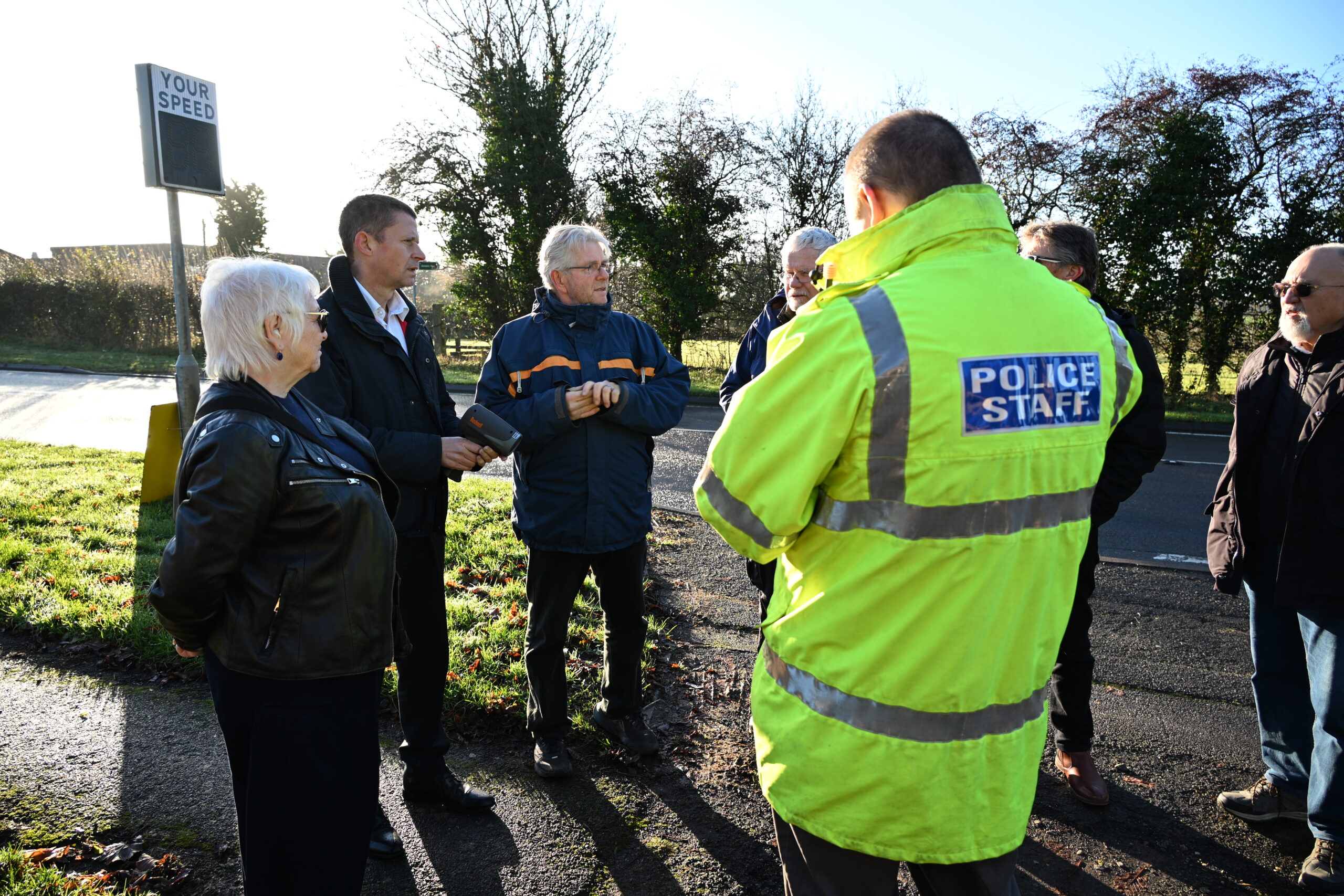 A group of people standing on a roadside in daylight, engaged in conversation. One person is wearing a bright yellow high-visibility jacket with reflective strips and the words “POLICE STAFF” printed on the back. Others are dressed in dark outdoor clothing. A speed monitoring sign is visible on the left, and the background shows trees and greenery under clear skies.