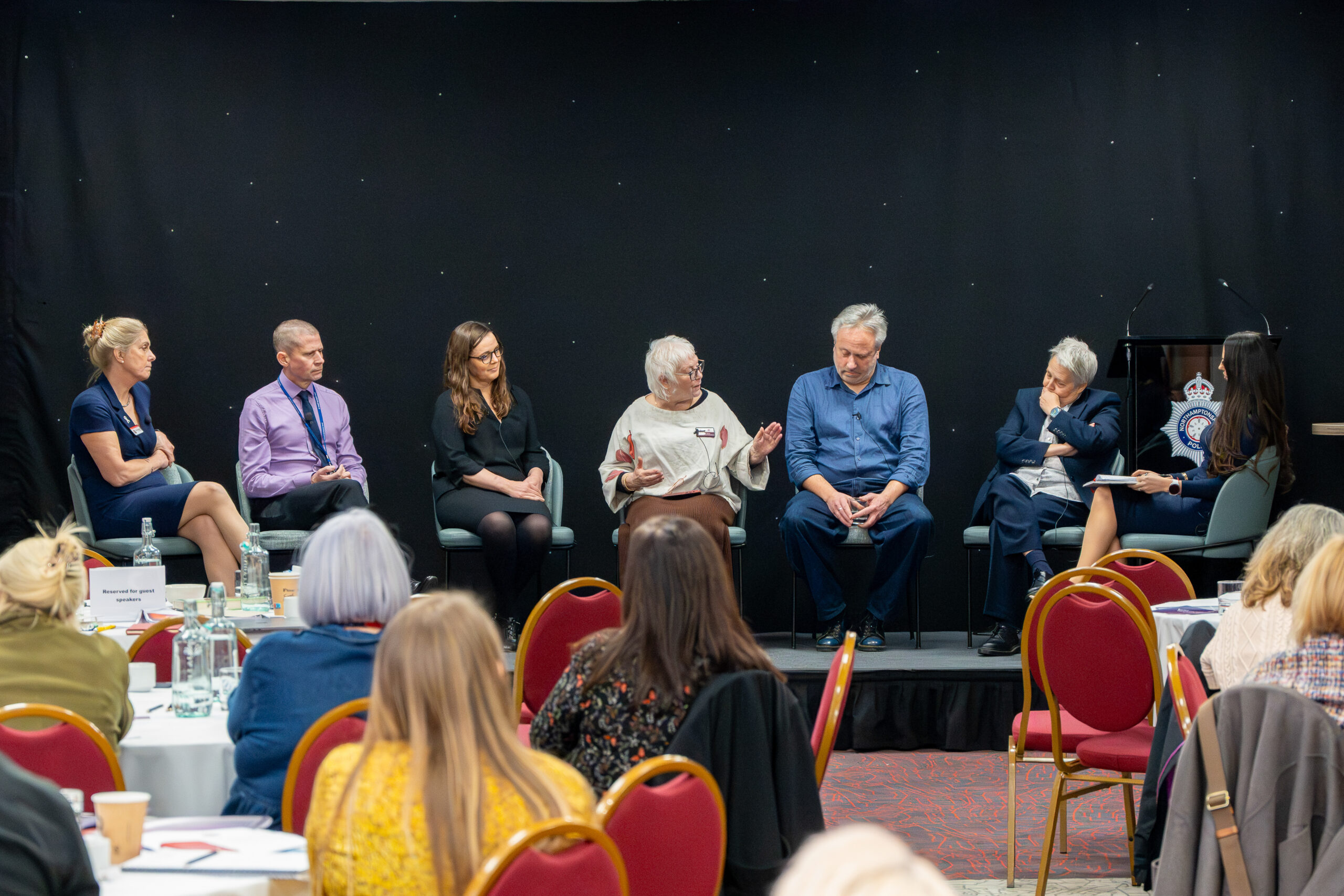 VAWG Conference A panel discussion taking place on a stage with six seated speakers and one moderator. The speakers are arranged in a row on chairs against a black backdrop, and the moderator is seated to the right near a lectern displaying a crest logo. In the foreground, several attendees are seated at round tables with white tablecloths, water bottles, and notepads. Red chairs with gold frames are visible throughout the room, and the setting appears to be a conference or seminar.
