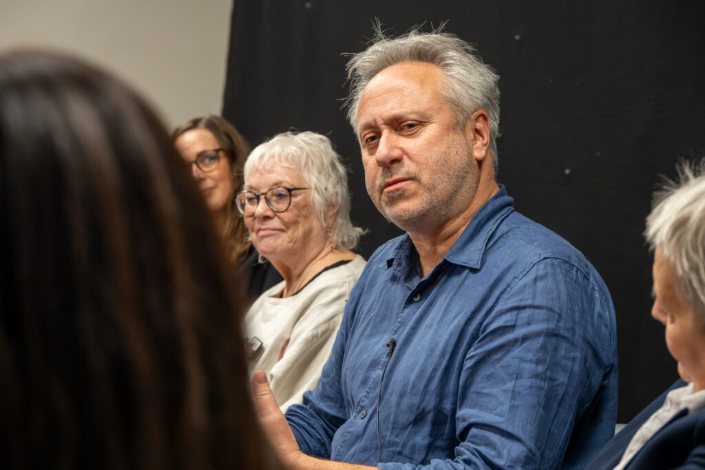 Close-up of a panel discussion showing several seated participants. The person in the foreground is wearing a blue long-sleeved shirt and appears to be speaking, with a microphone clipped to the shirt. Other participants are partially visible in the background, seated against a dark backdrop.