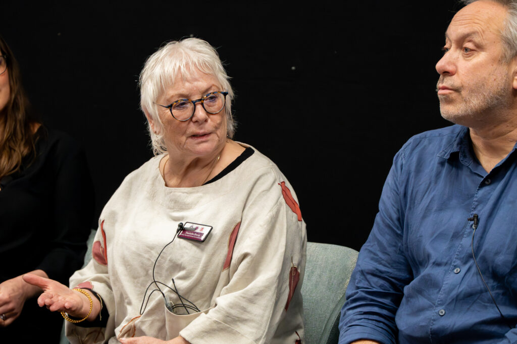 A close-up of three people seated during a panel discussion. The person in the center is wearing a light-colored top with a bird design and a name badge, and has a microphone wire clipped to the clothing. The person on the right is wearing a blue shirt, and the person on the left is partially visible in dark clothing. The background is plain and dark, suggesting an indoor conference setting.