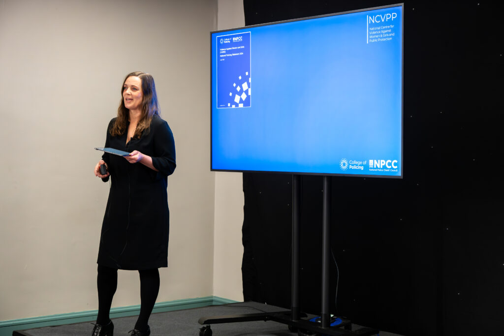 A person stands next to a large digital screen during a presentation. The screen displays a blue background with text and logos, including “NCVPP” at the top right and “College of Policing” and “NPCC” at the bottom right. The individual is holding a tablet and wearing a black outfit. The setting appears to be a conference or seminar room with a dark backdrop behind the screen.