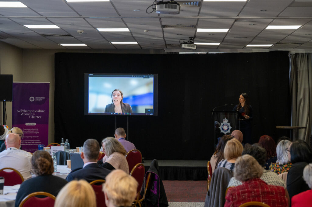 A conference setting with attendees seated at round tables facing a stage. On the stage, a large screen displays a video call participant, and a speaker stands at a lectern with a crest logo. To the left of the stage, a purple banner reads “Northamptonshire Women’s Charter” along with additional text. The room has a grid-patterned ceiling with lights and a projector, and red chairs with gold frames are arranged throughout.