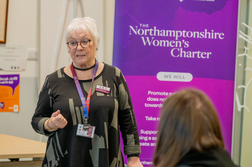 A person is standing and speaking in front of a purple banner that reads “The Northamptonshire Women’s Charter.” The banner includes text about promoting equality and supporting women’s voices. The person is wearing a dark patterned top and a lanyard with an ID badge. Another individual is seated in the foreground, facing the speaker.