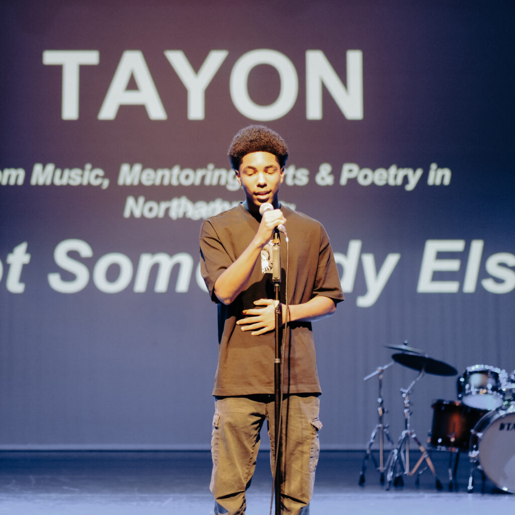 A person stands on a stage holding a microphone, lit by a spotlight. Behind them, a large screen displays the word “TAYON” along with additional text related to music, mentoring, arts, and poetry. A drum kit is visible in the background on the right side of the stage.