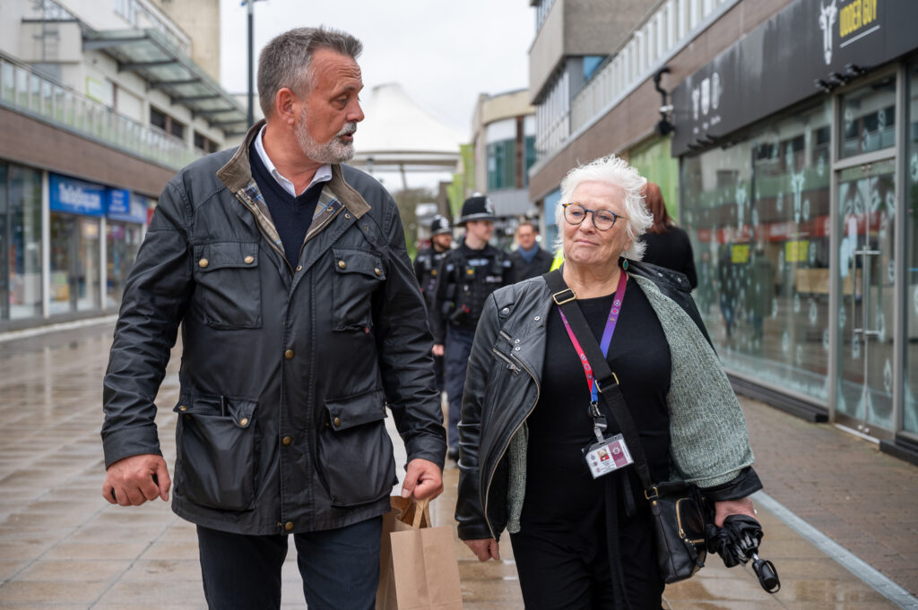 Two adults walk together along a pedestrian shopping street on a wet day. One carries a shopping bag, while the other wears a lanyard and holds a folded umbrella. Several police officers walk in the background, and shopfronts line both sides of the walkway.